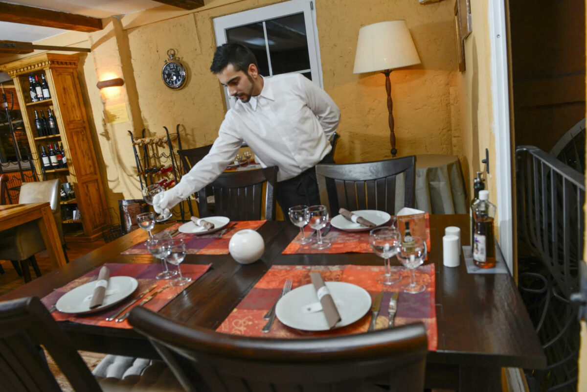 Kellner in italienischem Restaurant deckt einen Tisch für vier Personen; rustikales Ambiente mit Weindekor und gemütlicher Beleuchtung