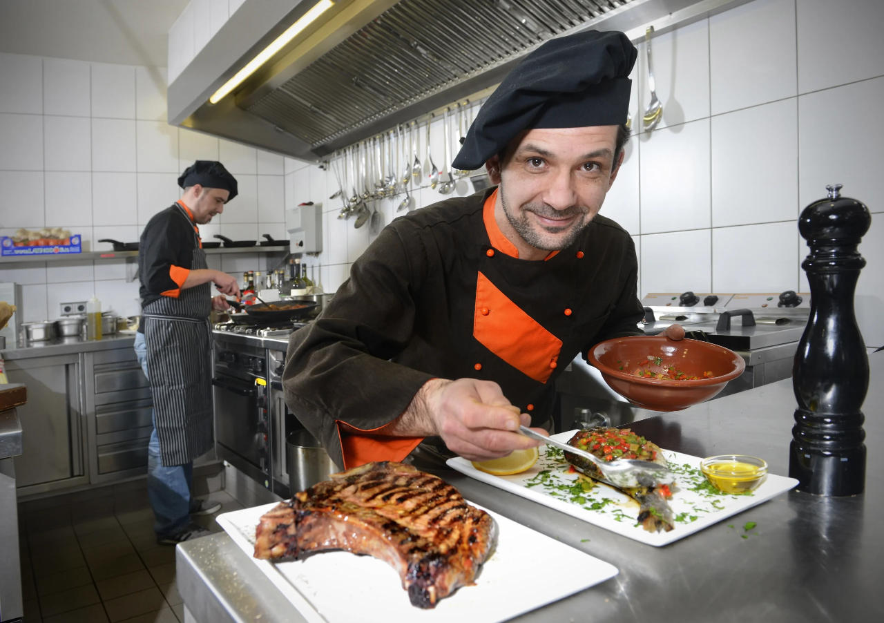 Koch in Restaurantküche präsentiert mediterrane Gerichte wie gegrillten Fisch und Steak; zweiter Koch arbeitet im Hintergrund